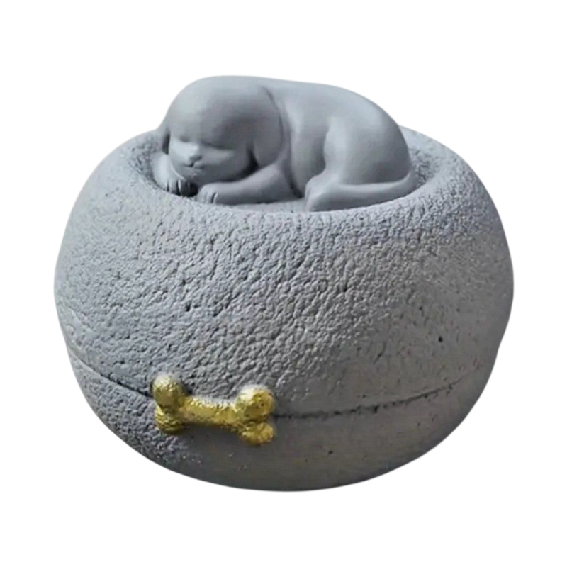 Grijze honden urn: een vredig slapend hondje bovenop een ronde, steenachtige basis, met een subtiel gouden botje als detail.