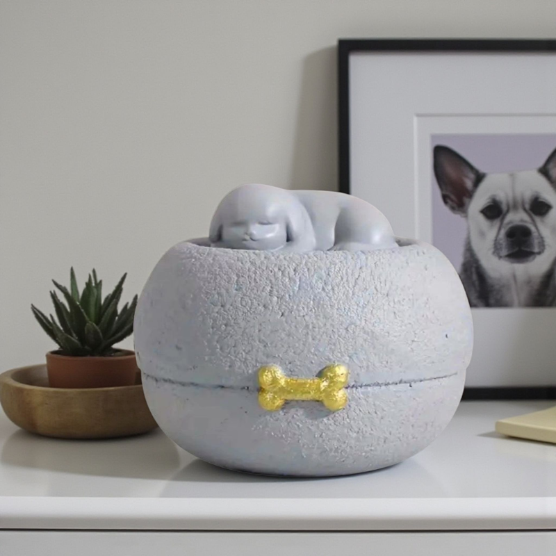 Grijze honden urn: een vredig slapend hondje bovenop een ronde, steenachtige basis, met een subtiel gouden botje als detail.