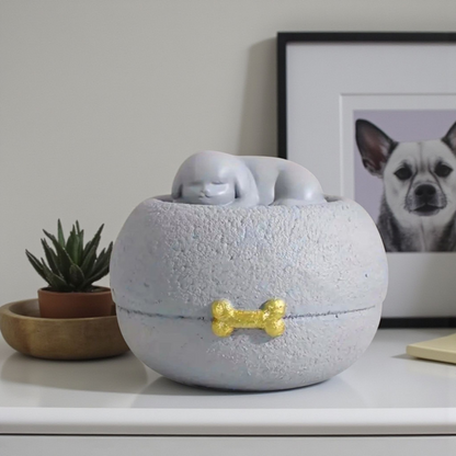 Grijze honden urn: een vredig slapend hondje bovenop een ronde, steenachtige basis, met een subtiel gouden botje als detail.