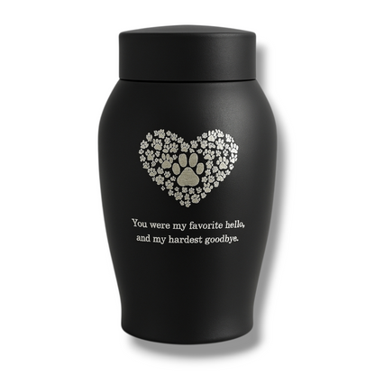 Zwarte matte urn met hart van pootafdrukken en troostende tekst, gefotografeerd tegen een lichte achtergrond.

